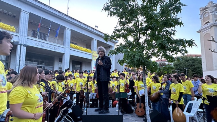 Άνδρας με μικρόφωνο πάνω σε βάθρο, ανάμεσα από παιδιά στα κίτρινα με μουσικά όργανα