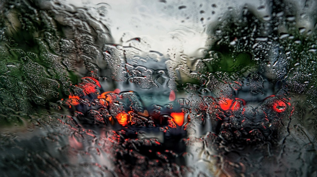 Τζάμι αυτοκινήτου γεμάτο σταγόνες βροχής και μπροστά τα φώτα από αυτοκίνητα