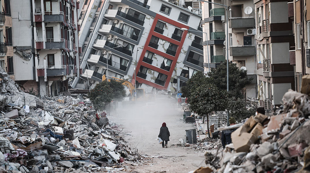 Γυναίκα στην πόλη Χατάι περπατά μπροστά από σπίτι που έχει γείρει μετά τον καταστροφικό σεισμό στην Τουρκία