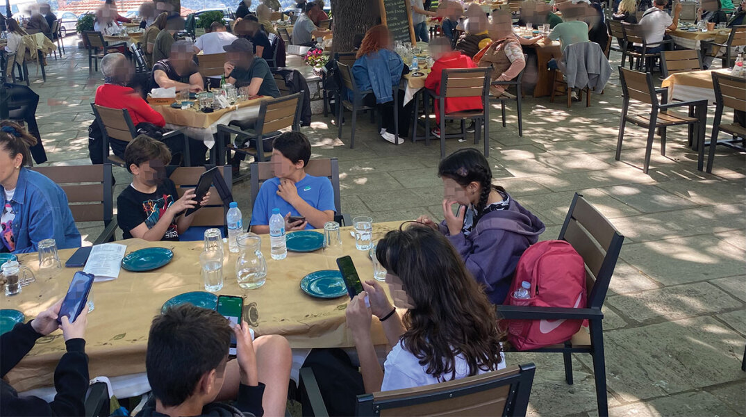 Παιδιά καθισμένα σε τραπέζι ταβέρνας με τα κινητά στο χέρι