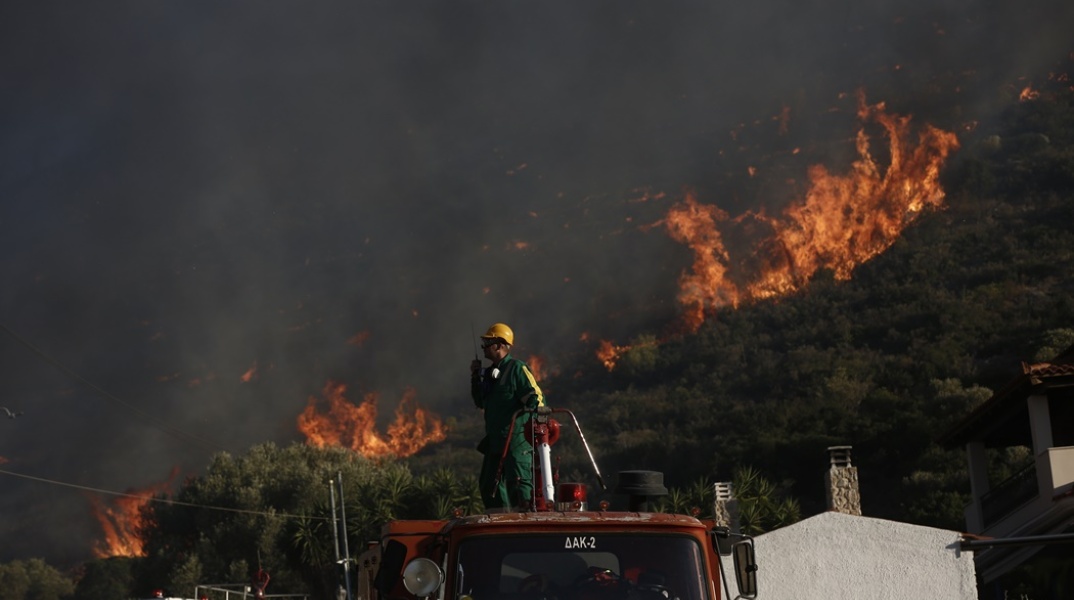 Πυροσβέστης πάνω στο όχημα δίνει οδηγίες μέσω ασυρμάτου - Από πίσω του οι φωτιές στον Κουβαρά