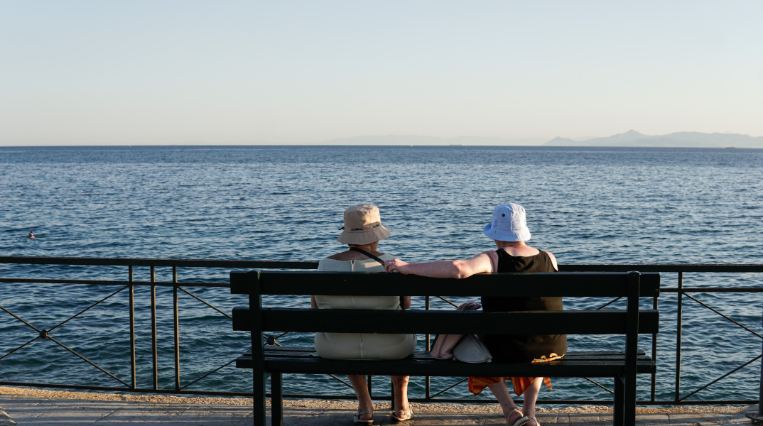 Γυναίκες σε παγκάκι απολαμβάνουν τη θέα προς τη θάλασσα και φορούν καπέλο για την προστασία τους από τον ήλιο