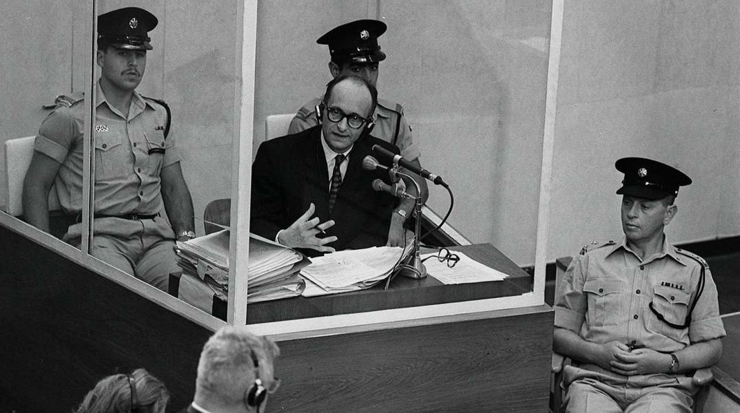 Ο Ναζί εγκληματίας πολέμου Άντολφ Άιχμαν στέκεται σε προστατευτικό γυάλινο θάλαμο πλαισιωμένο από την ισραηλινή αστυνομία κατά τη διάρκεια της δίκης του στις 22 Ιουνίου 1961 στην Ιερουσαλήμ