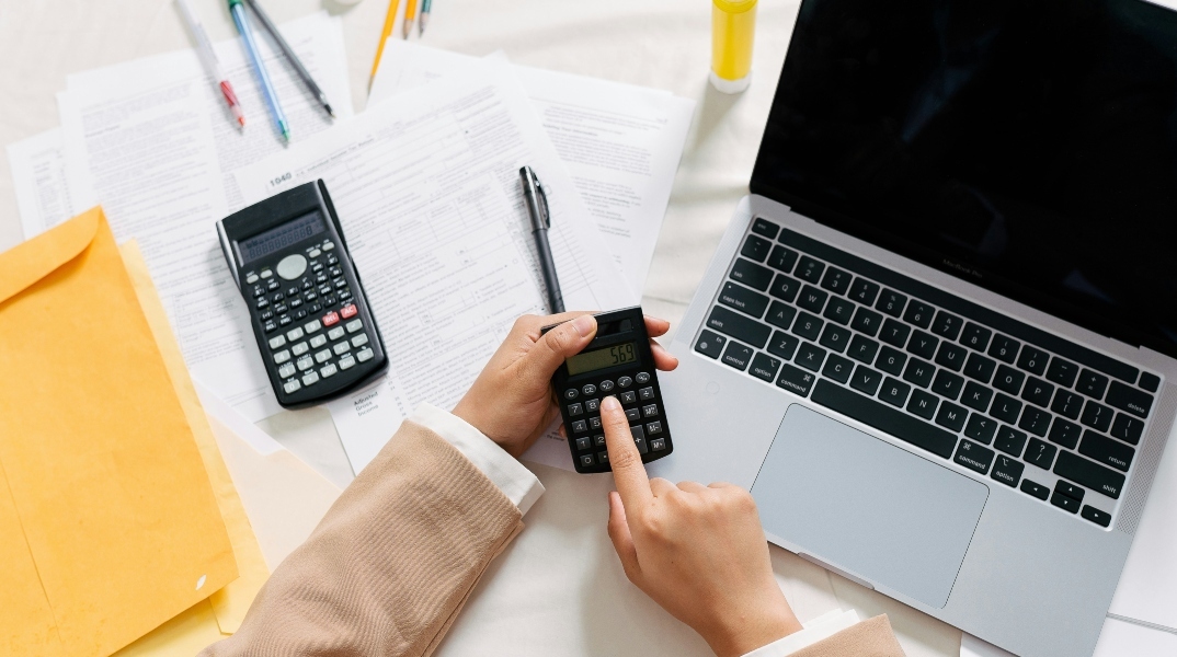 Δύο χέρια χειρίζονται calculator πάνω από ένα laptop μαζί με λογιστικά έγγραφα