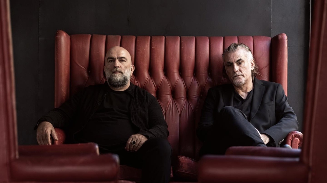 Ο Φίλιππος Πλιάτσικας και ο Μπάμπης Στόκας καθισμένοι σε κόκκινο καναμπέ φορώντας μαύρα ρούχα, κοιτούν το φακό