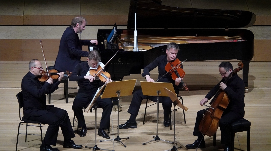 4 μουσικοί με έγχορδα, βιολί - βιολοντζέλο σε παράσταση. Από πίσω τους ένα πιανίστας παίζε με μαύρο πιάνο με ουρά