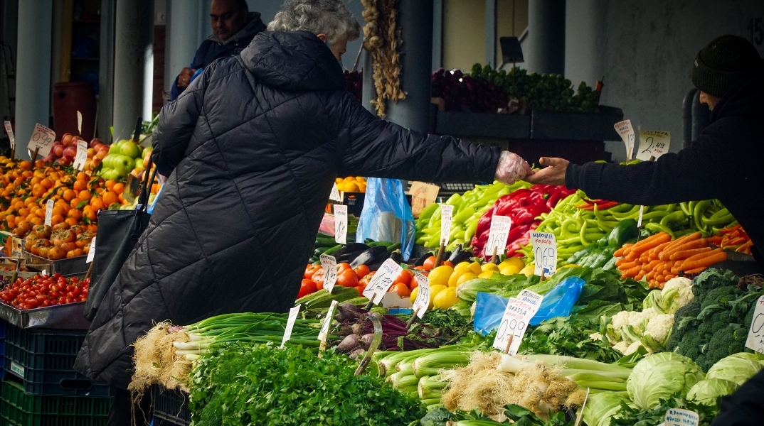 Γυναίκα όρθια με μαύρο παλτό δίνει λεφτά σε άνδρα με μαύρο σκούφο πάνω από τελάρα με λαχανικά