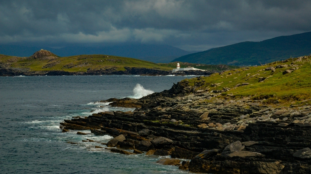 Βραχώδης παραλία και στο βάθος άσπρο κτίριο και κόκκινος φάρος