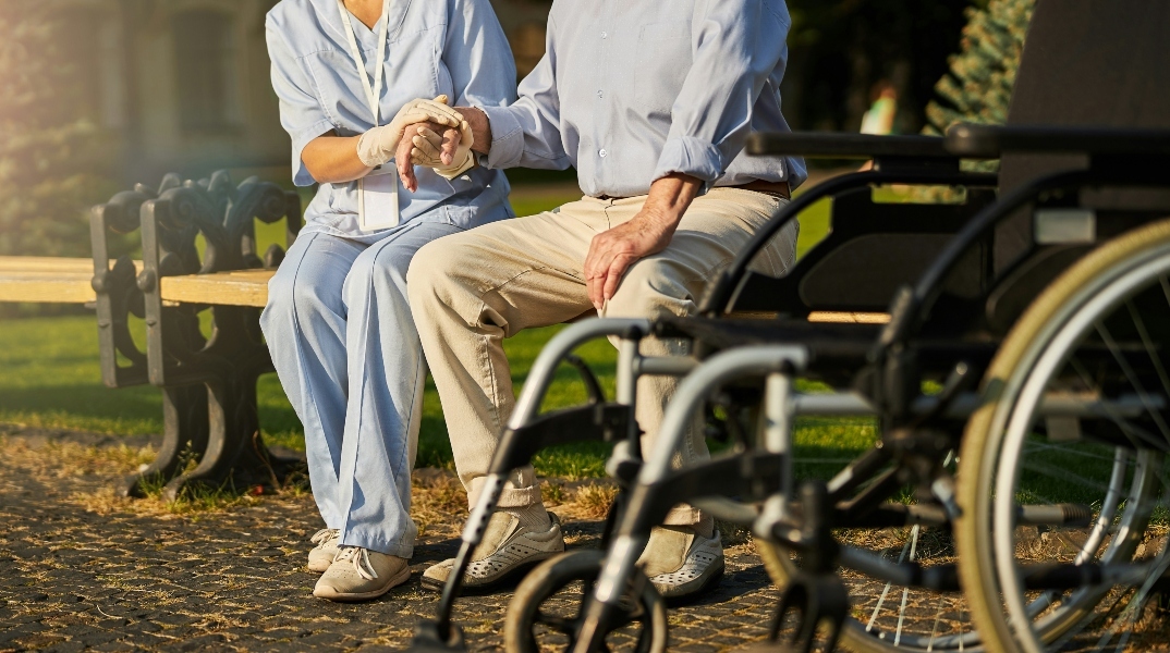 Νοσοκόμα κρατάει το χέρι ηλικιωμένο - κάθονται σε παγκάκι πίσω από αναπηρική καρέκλα
