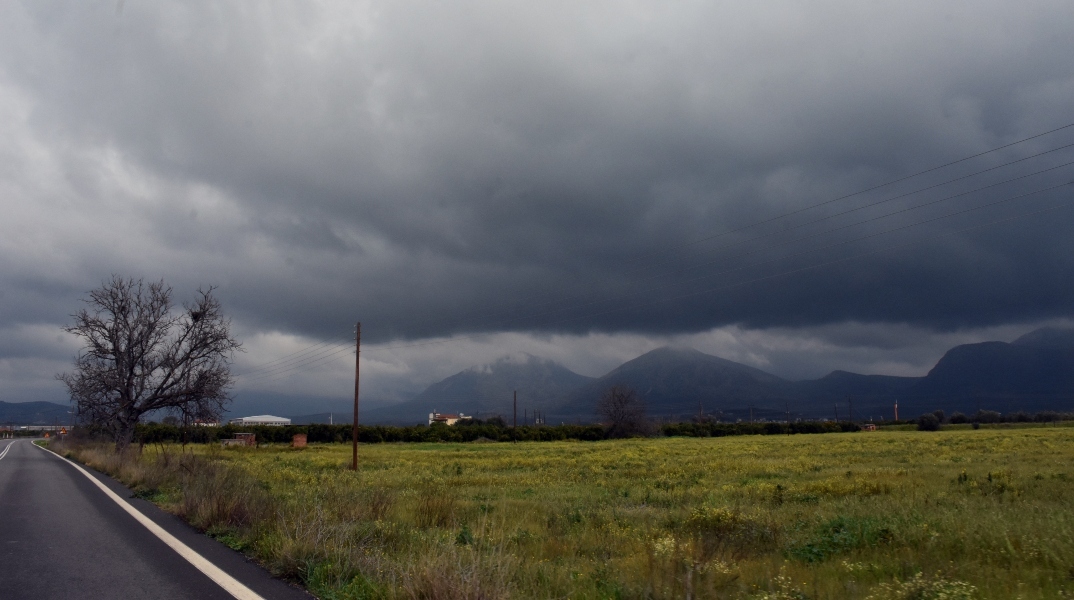 Επαρχιακός δρόμος δίπλα σε πράσινο χωράφι - Στο βάθος βουνά και σύννεφα