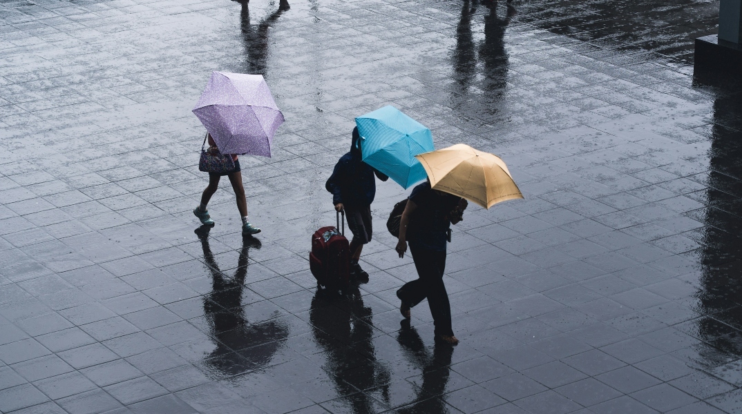 Μία γυναίκα και δύο άνδρες περπατούν στην βροχή κρατώντας ομπρέλες
