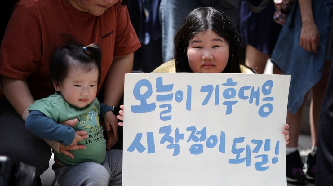 Νεαρή Κορεατισσα κρατάει πινακίδα δίπλα σε γονέα με ένα μωρό