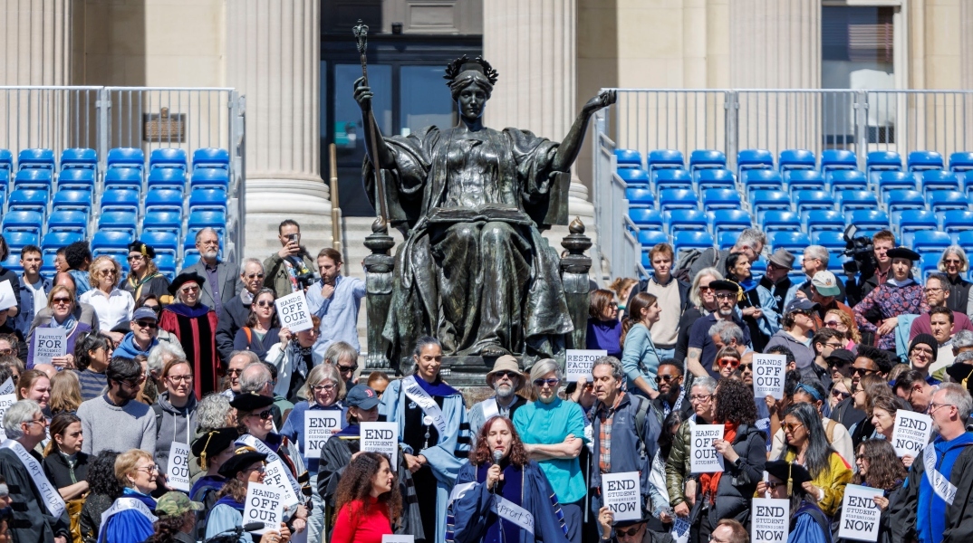 Καθηγητές ντυμένοι με παραδοσιακές ρόμπες μπροστά από άγαλμα κρατούν πλακάτ