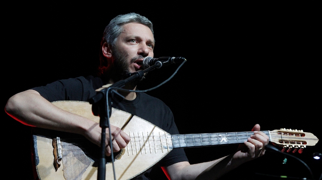 Ο Αλκίνοος Ιωαννίδης καθιστός μπροστά σε μικρόφωνο τραγουδάει κρατώντας λάουτο