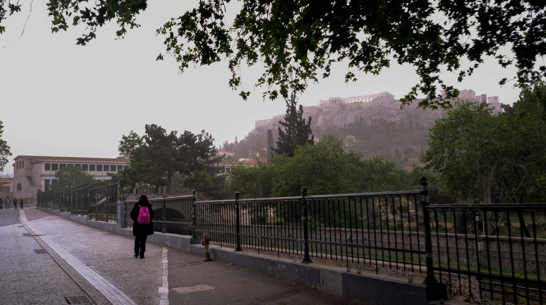 Άνθρωπος στα μαύρα περπατά σε δρόμο δίπλα σε κάγκελα με κόκκινη τσάντα