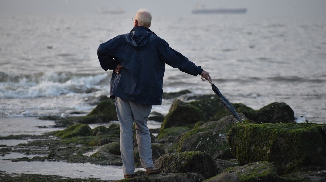 Ηλικιωμένος με ομπρέλα στην ακτή με φόντο τη θάλασσα