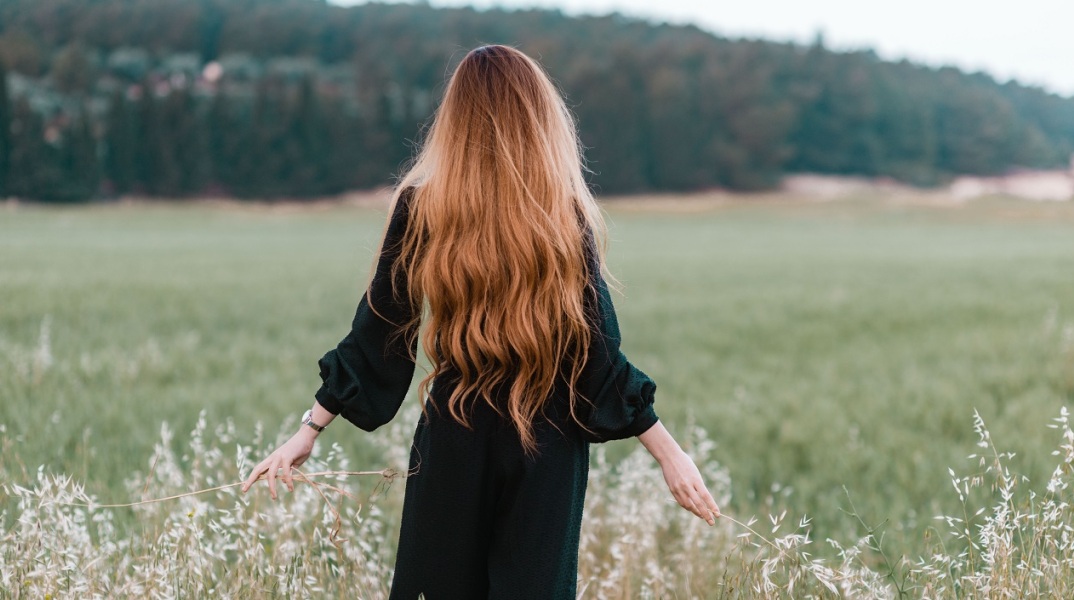 Κοπέλα με μακριά μαλλιά και γυρισμένη πλάτη περπατάει σε χωράφι αγγίζοντας τα φυτά