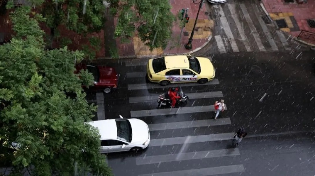 Πολίτες περπατούν υπό βροχή ανάμεσα σε αυτοκίνητα και μηχανάκι στο κέντρο της Αθήνας