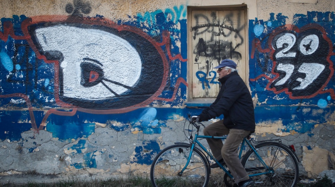 Ηλικιωμένος ποδηλάτης περνά μπροστά από γκράφιτι σε τοίχο εγκαταλειμμένου σπιτιού 