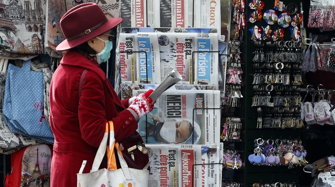 Γυναίκα που φορά ιατρική μάσκα προστασίας στέκεται μπροστά σε περίπτερο με γαλλικές εφημερίδες