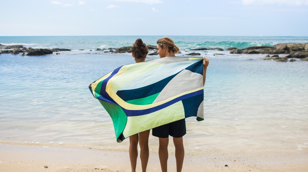 Άντρας και γυναίκα στέκονται με γυρισμένη πλάτη όρθιοι σε παραλία, τυλιγμένοι με μια πετσέτα