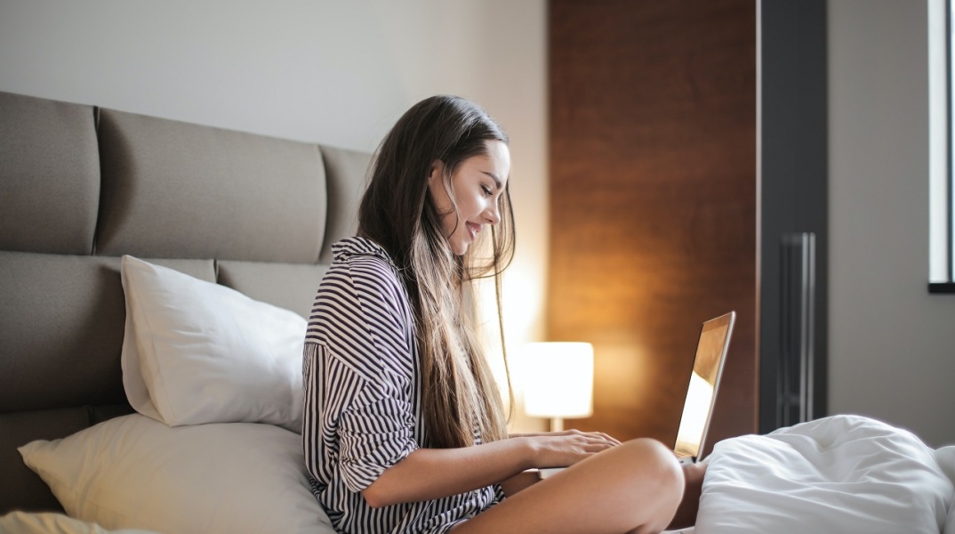 Κοπέλα καθισμένη στο κρεβάτι με ανοιχτό laptop μπροστά της