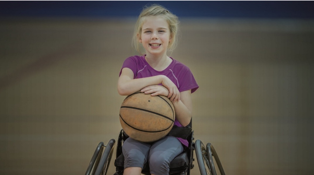 Κοριτσάκι καθισμένο σε αναπηρικό καρότσι χαμογελάει κρατώντας μια μπάλα μπάσκετ στα χέρια της