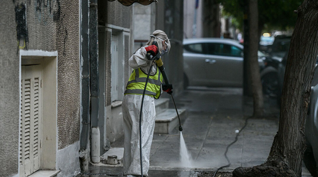 Υπάλληλος του Δήμου Αθηναίων με προστατευτική στολή για τον κορωνοϊό απολυμαίνει πεζοδρόμιο στο κέντρο της Αθήνας