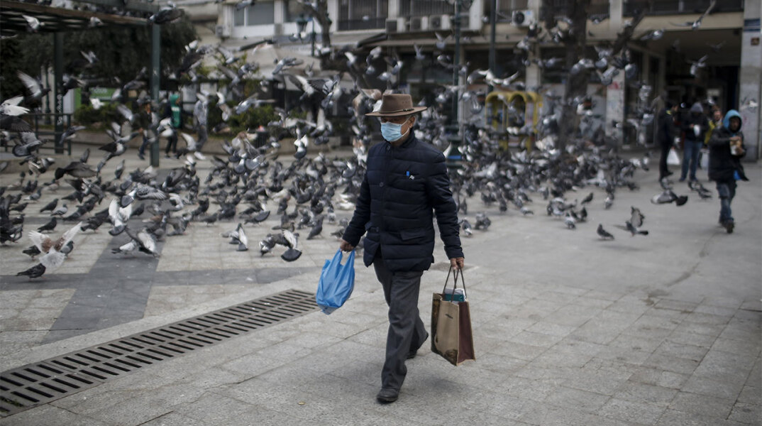Ηλικιωμένος άνδρας με προστατευτική μάσκα για τον κορωνοϊό περπατά στο κέντρο της Αθήνας, κουβαλώντας τσάντες με ψώνια