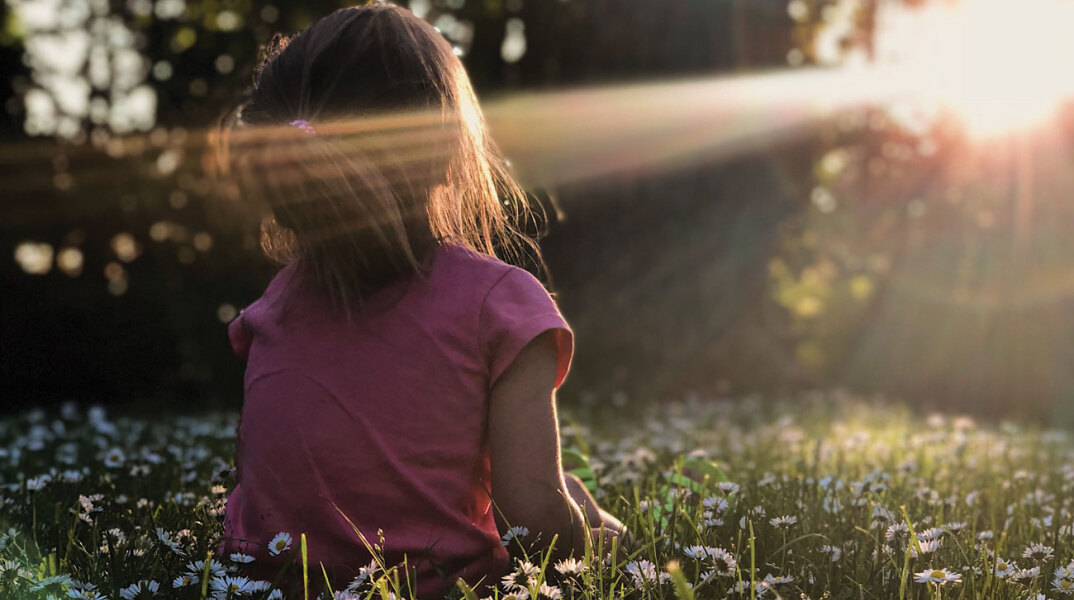 Κοριτσάκι γυρισμένο πλάτη κάθεται στο γρασίδι με φόντο δέντρα και ήλιο