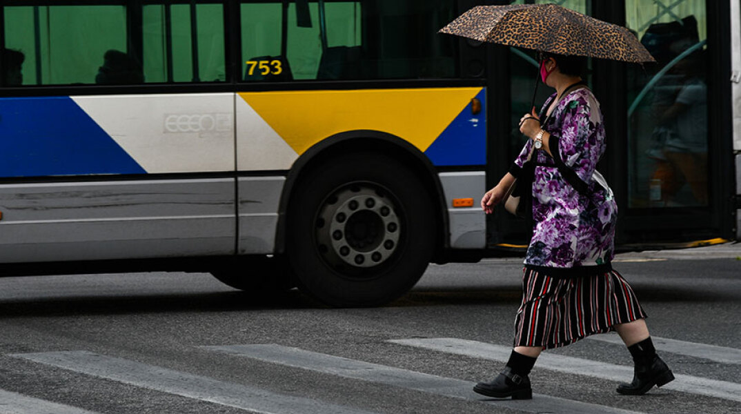 Γυναίκα διασχίζει διάβαση πεζών στον Πειραιά, κρατώντας ομπρέλα και φορώντας προστατευτική μάσκα για τον κορωνοϊό