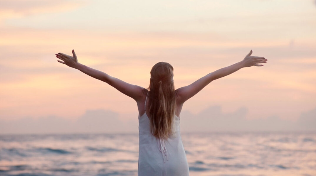 Κοπέλα με γυρισμένη πλάτη και ανοιχτά χέρια με φόντο τον ουρανό
