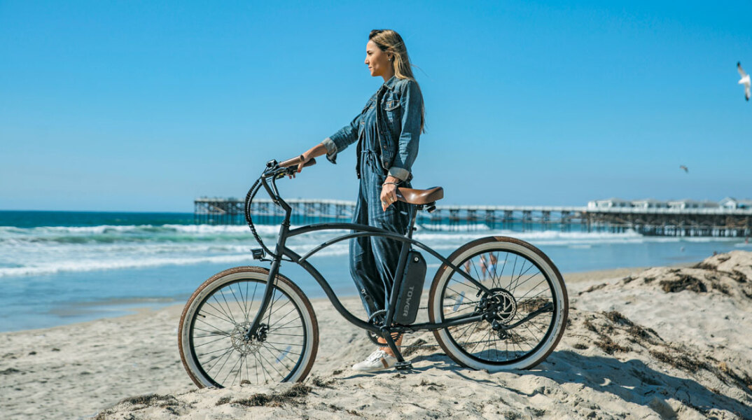 Κοπέλα στην άμμο, δίπλα στη θάλασσα, στέκεται δίπλα σε ένα ποδήλατο κρατώντας το τιμόνι του