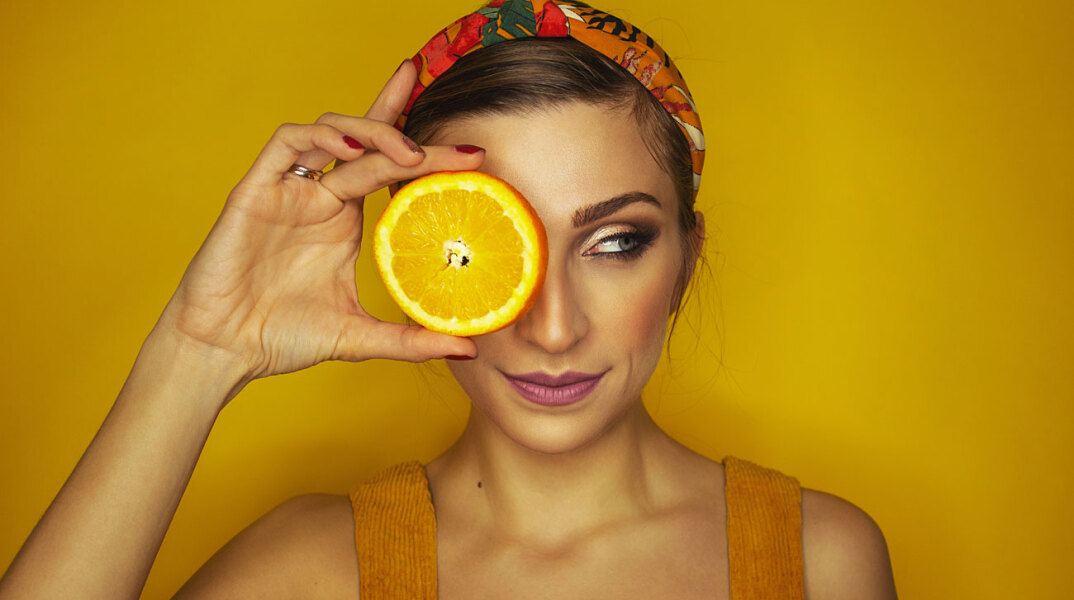 Καστανή κοπέλα με μαζεμένα μαλλιά κρατάει μια φέτα πορτοκάλι μπροστά από το αριστερό της μάτι, σε πορτοκαλί φόντο