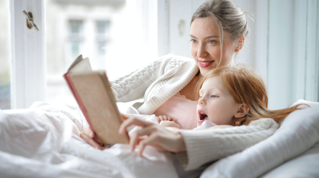 Μητέρα και κόρη ξαπλωμένες στον καναπέ αγκαλιά με ανοιχτό βιβλίο