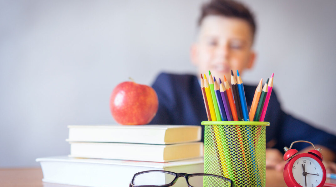 Βιβλία, μήλο, μολύβια ακουμπισμένα σε γραφείο και στο βάθος αγόρι καθισμένο