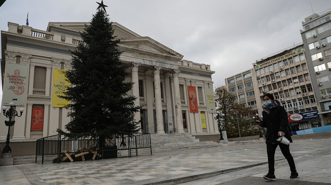 Γυναίκα με προστατευτική μάσκα περνά μπροστά από το Δημοτικό Θέατρο Πειραιά, όπου έχει στηθεί ένα τεράστιο χριστουγεννιάτικο δέντρο