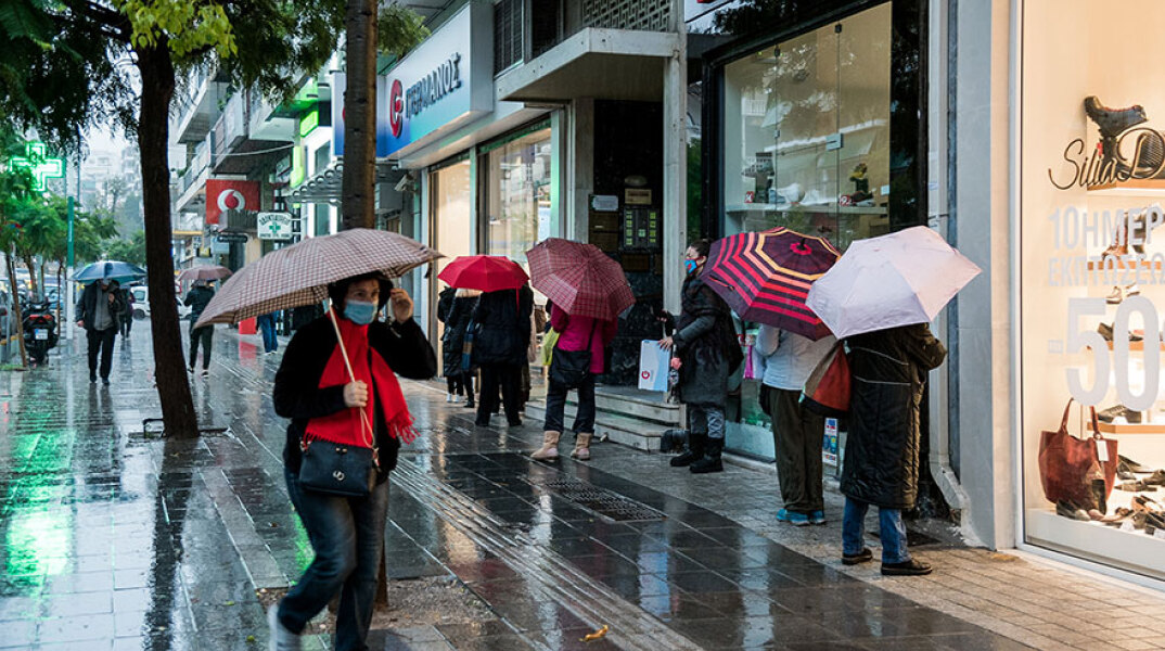Πολίτες με ομπρέλες και μάσκες για τον κορωνοϊό σε πεζόδρομο εν μέσω βροχής