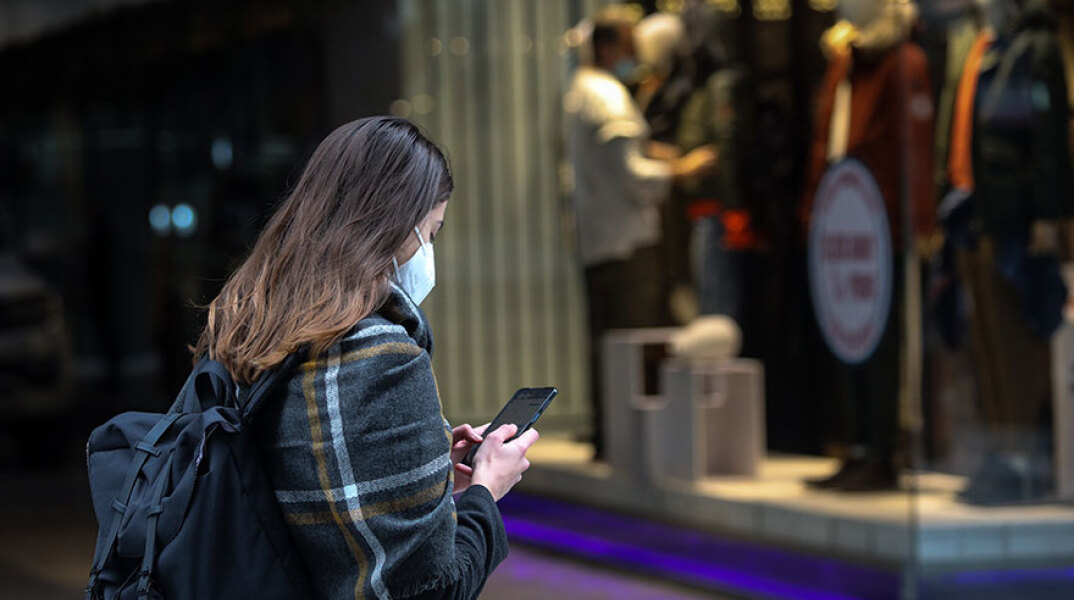 Κοπέλα με μάσκα για τον κορωνοϊό κοιτάζει το κινητό της με φόντο καταστήματα στην Ερμού
