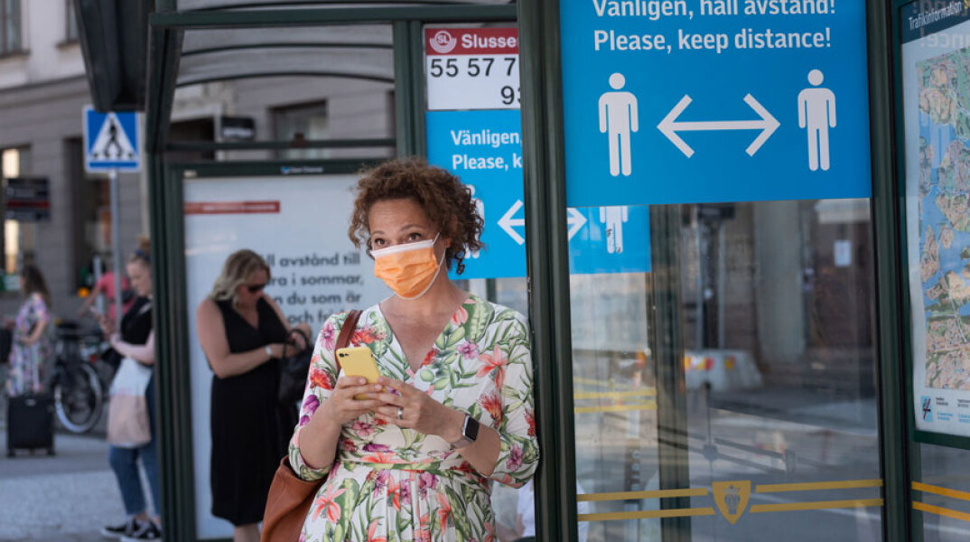 Κορωνοϊός: Κοπέλα με μάσκα και το κινητό στο χέρι περιμένει σε στάση λεωφορείου