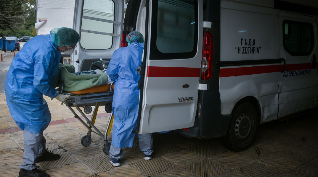 Υγειονομικοί με προστατευτικές στολές για τον κορωνοϊό φορτώνουν φορείο σε ασθενοφόρο του ΕΚΑΒ