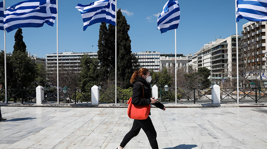 Γυναίκα με μάσκα για τον κορωνοϊό περνά από το Σύνταγμα όπου έχουν τοποθετηθεί ελληνικές σημαίες για τα 200 χρόνια από την Επανάσταση του 1821