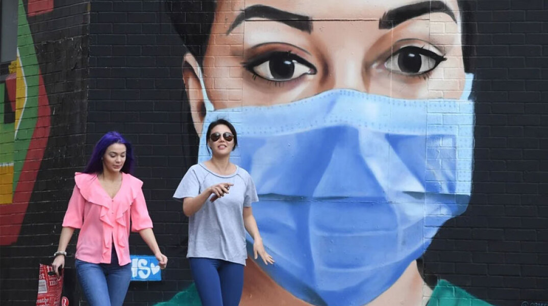 Νεαρές κοπέλες χωρίς μάσκα περπατούν στο Λονδίνο με φόντο ένα γκράφιτι για την πανδημία Covid-19