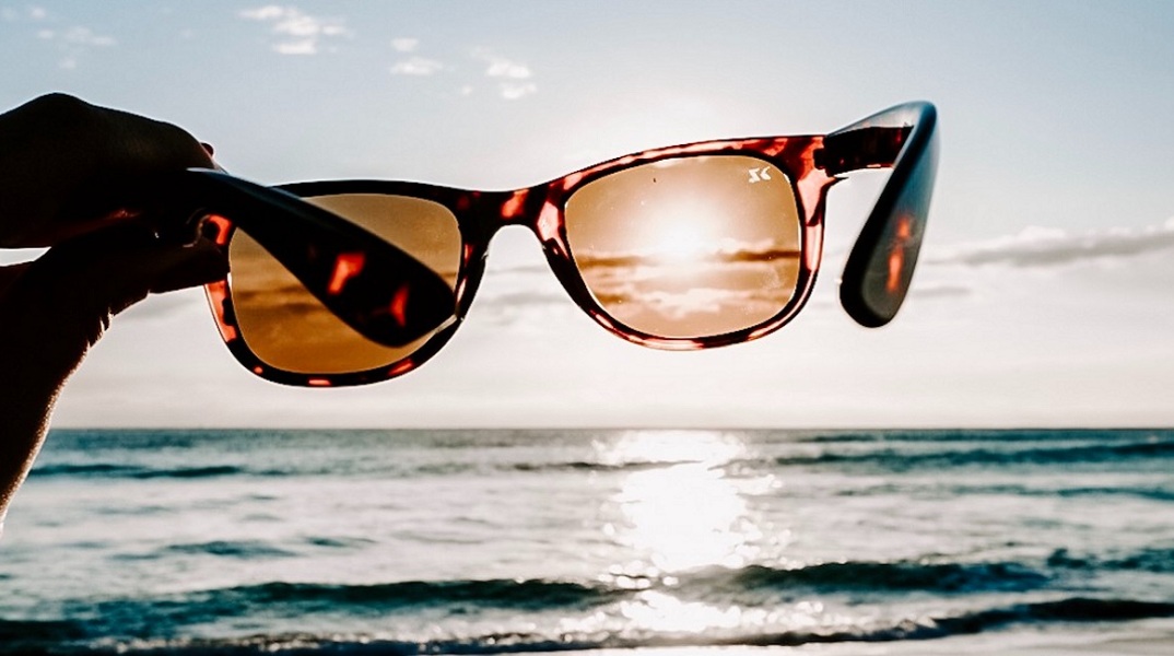 Ένα άτομο κρατάει ένα ζευγάρι γυαλιά ηλίου μπροστά από τη θάλασσα. 