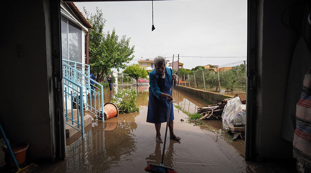 Πλημμύρες στη Βόρεια Εύβοια: Ηλικιωμένη με σκούπα απομακρύνει το νερό της βροχής που έφερε λάσπες και χώματα στο σπίτι της