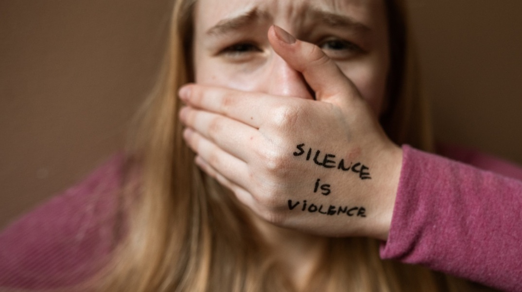 Μια γυναίκα που κλαίει, κλείνει το στόμα της και το χέρι της γράφει «η σιωπή είναι βία».