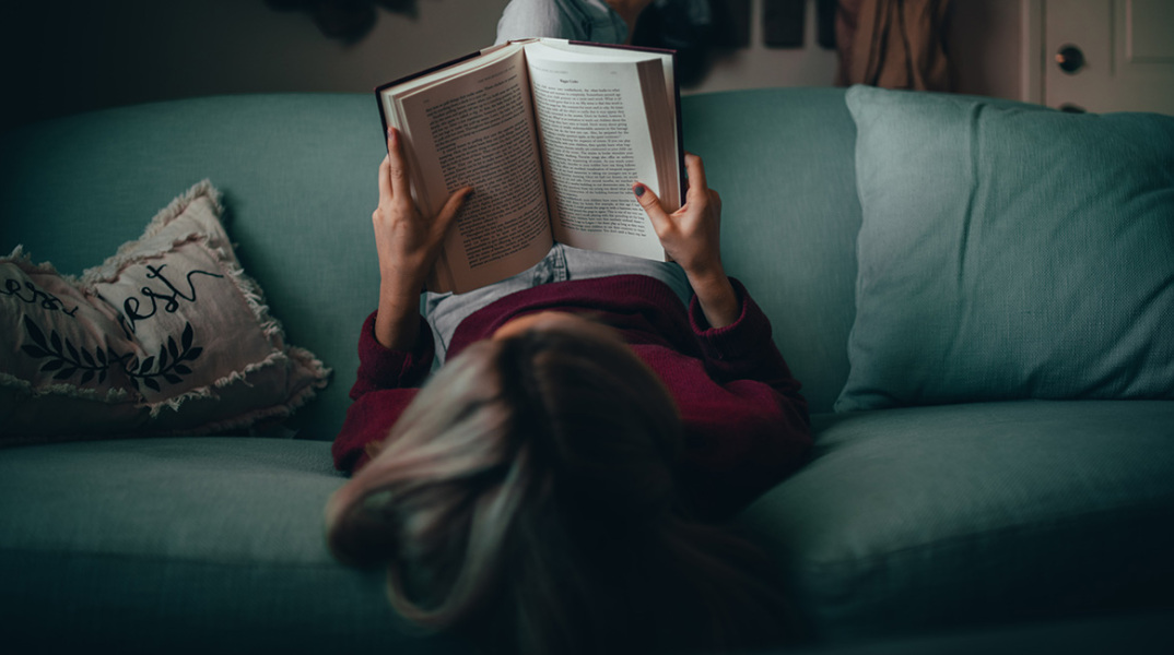 Μια κοπέλα κάθεται ανάποδα στον καναπέ και διαβάζει βιβλίο
