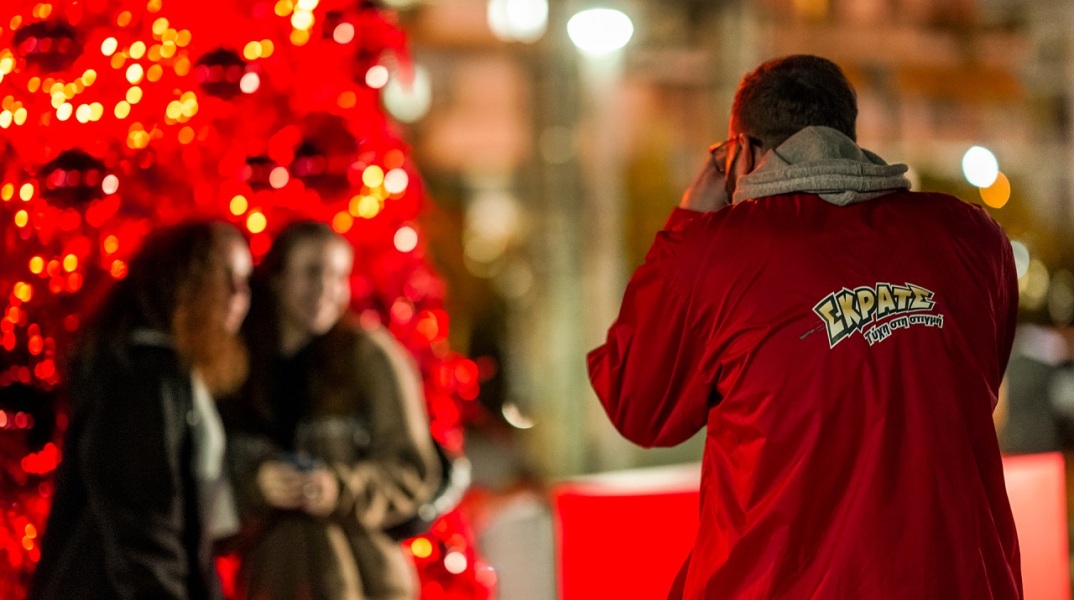 Άνδρας ντυμένος στα κόκκινο βγάζει φωτογραφία δύο κοπέλες μπροστά από ένα κόκκινο χριστουγεννιάτικο δέντρο
