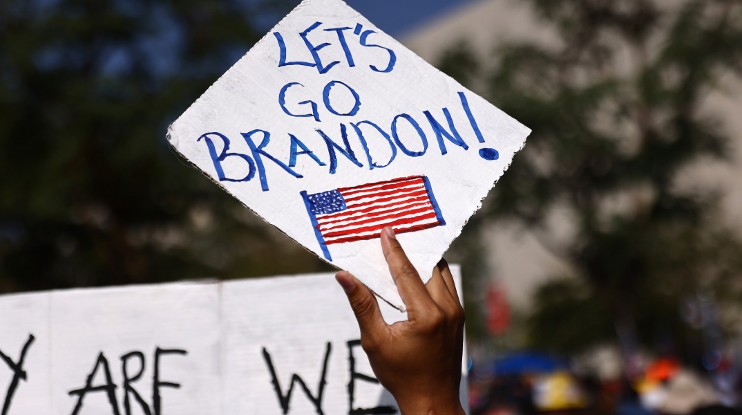 Χέρι που κρατά πινακίδα στην οποία αναγράφεται η φράση Let's Go Brandon