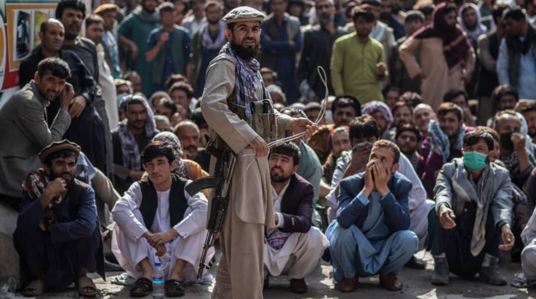 Ένας φρουρός ασφαλείας των Ταλιμπάν κρατάει μαστίγιο ενώ στέκεται μπροστά σε Αφγανούς άνδρες που περιμένουν έξω από μια τράπεζα για να βγάλουν χρήματα 